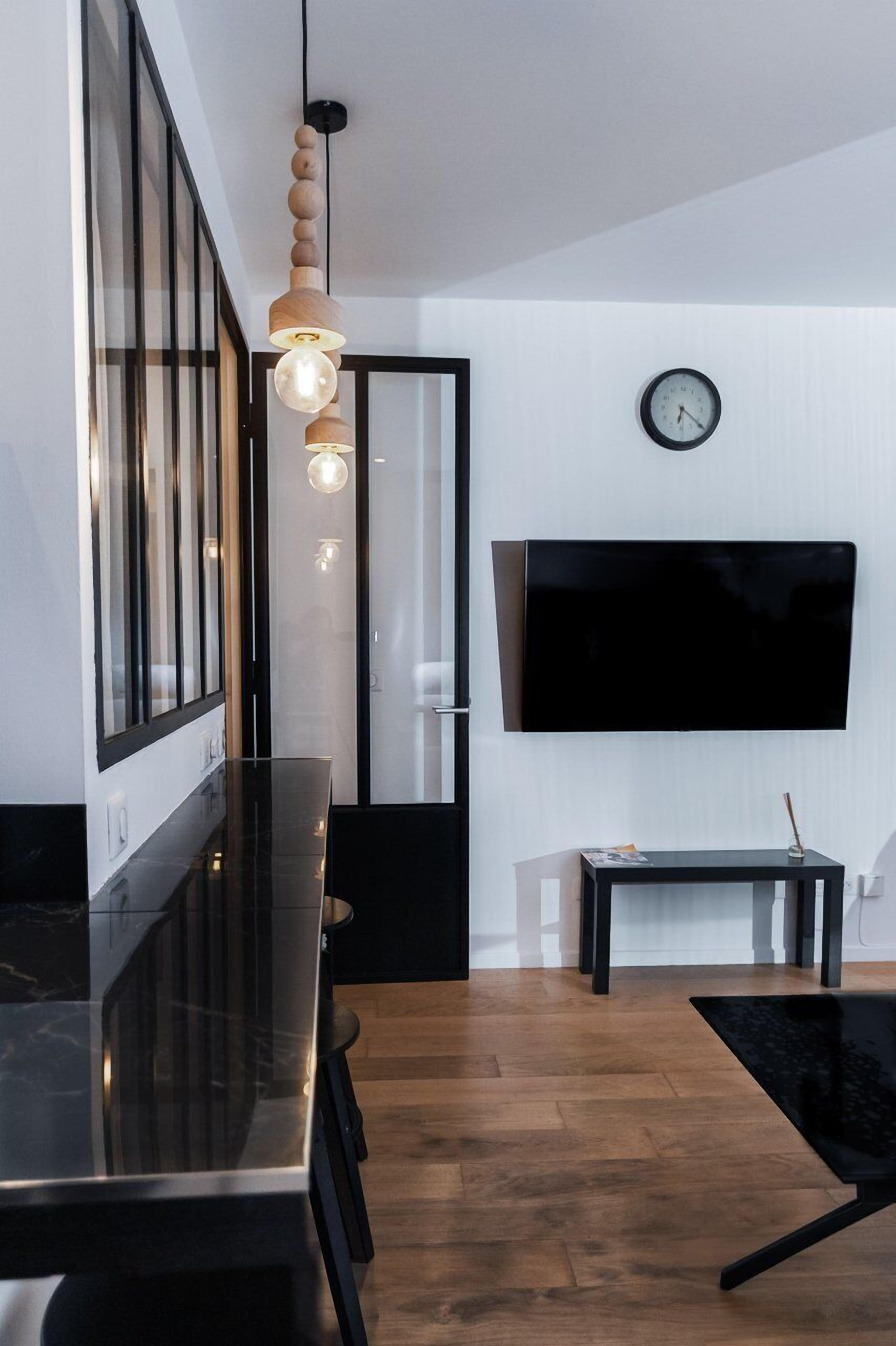 A modern living room with a black sofa, a glass coffee table, a wooden pendant light, a flat-screen TV, and a clock on the wall.