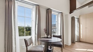 Een moderne kamer met een ronde tafel, twee stoelen, een vaas met bloemen en grote ramen voorzien van gordijnen.