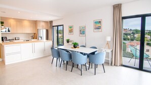 Eine moderne Küche mit weißen Schränken, ein Essbereich mit weißem Tisch und blauen Stühlen sowie ein Balkon mit Blick auf die Stadt.