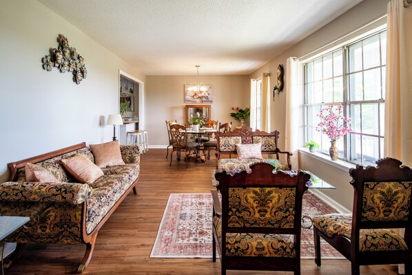 A spacious living room with a wooden floor, a sofa, chairs, a dining table, and a large window with a view.