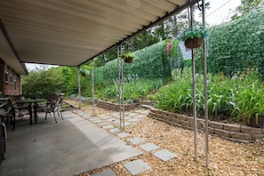 A covered patio with a table and chairs, surrounded by a garden with various plants and a stone pathway.