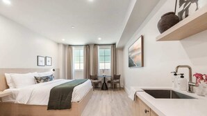 Una habitación de hotel moderna con una cama, una pequeña mesa con sillas, un lavabo y un ventanal con cortinas.