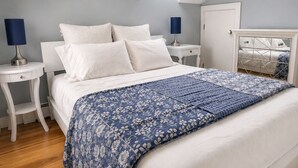 Un dormitorio con tragaluz, cama con ropa de cama blanca, manta estampada en azul y suelo de madera.