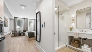 Een moderne hotelkamer met kitchenette, een bed, zithoek met tafel en stoelen, grote spiegel, douchecabine van glas en een witte badkamer met wastafel en handdoekenkast.