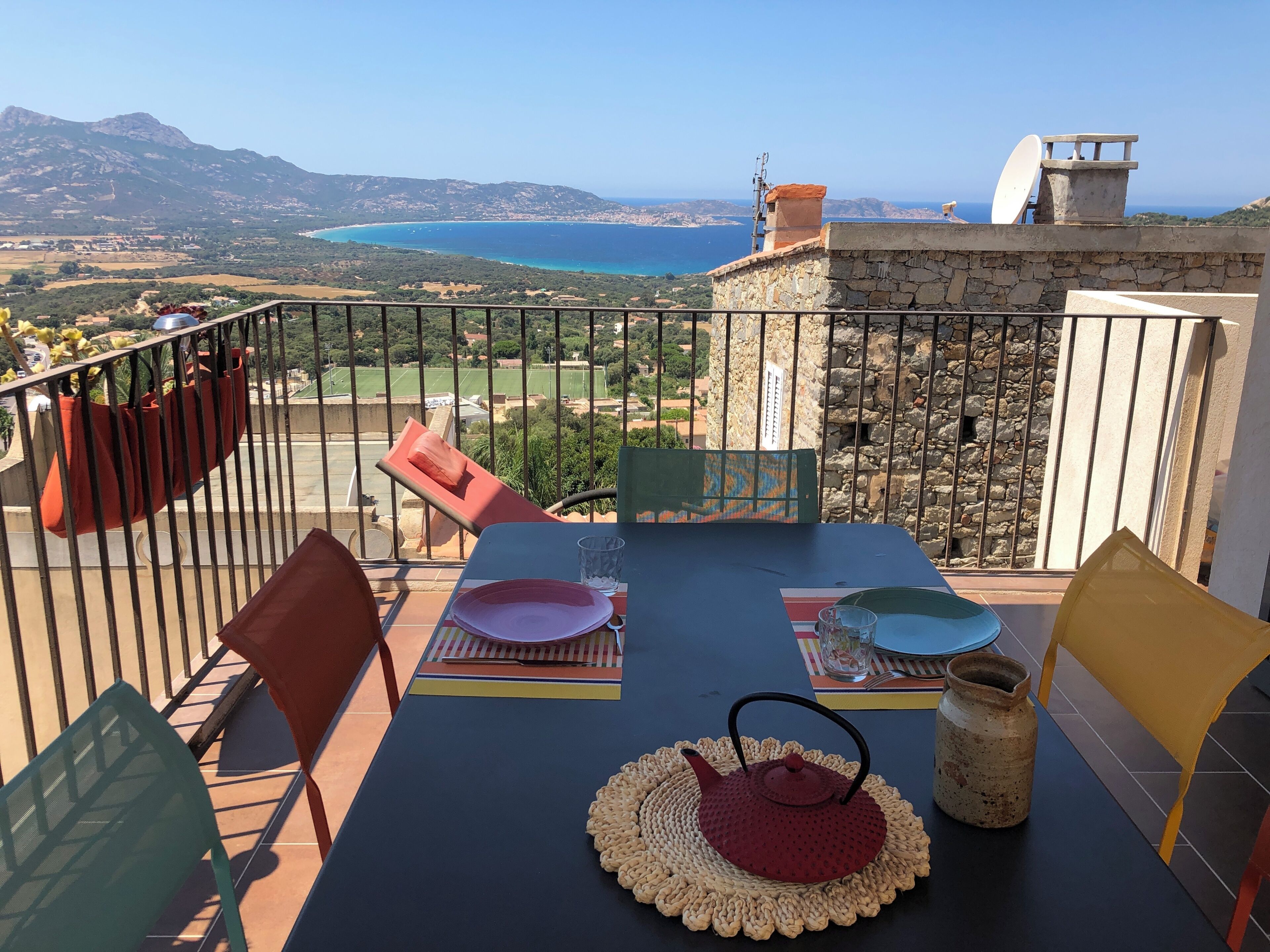 Un balcon avec une table dressée pour deux personnes, une bouilloire rouge et une vue sur la mer et les montagnes.