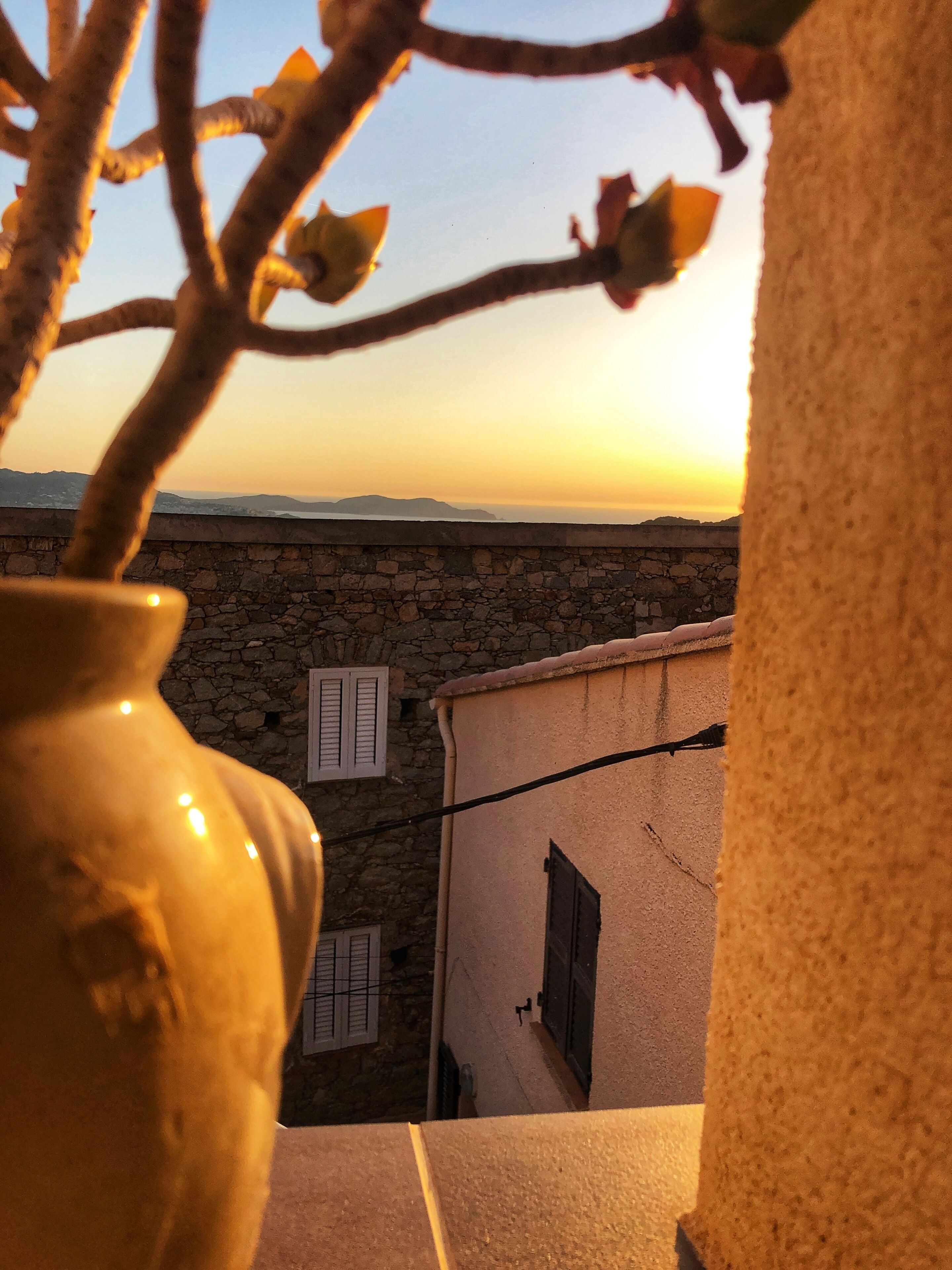Un mur de pierre avec des volets, un vase et une vue sur un coucher de soleil se dégageant sur un paysage lointain.