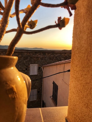 Eine Steinsäule mit Fensterläden, einer Vase und Blick auf einen Sonnenuntergang über einer entfernten Landschaft.