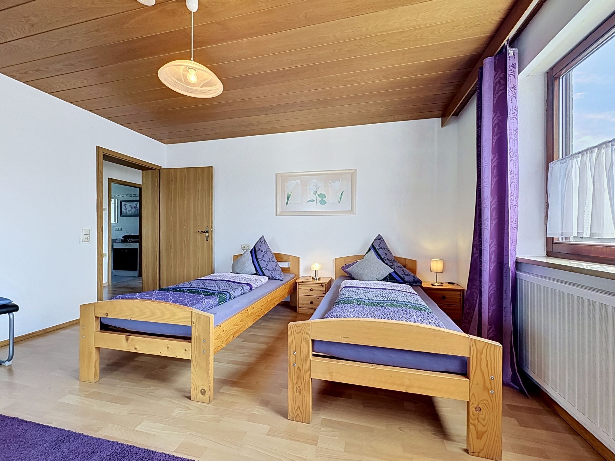 A room with two wooden beds, a ceiling light, a framed picture on the wall, and a window with curtains.