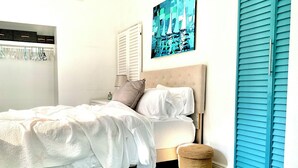 A bedroom with a bed, a bedside table, a lamp, a painting, a window with shutters, and a door with shutters. - Serene- pool house with large enclosed oasis of local artist in Port Charlotte (Port Charlotte)