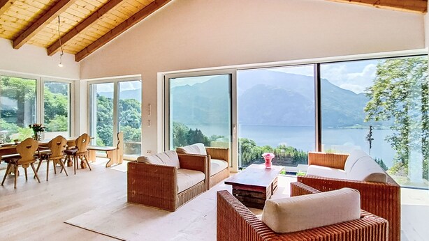 Una habitación amplia con ventanas grandes, vigas de madera en el techo, muebles de mimbre y vistas a un lago y montañas.