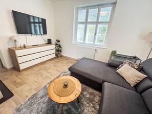 A modern living room with a grey sofa, a wooden coffee table, a TV mounted on the wall, and a window with blinds.