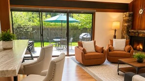 A modern living room with a fireplace, a dining area with a table and chairs, and a large sliding glass door leading to a backyard.
