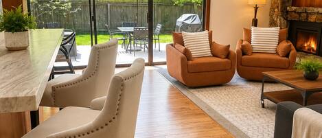 A modern living room with a fireplace, a dining area with a table and chairs, and a large sliding glass door leading to a backyard.