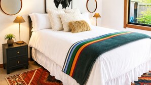 A bedroom with a bed, two bedside tables, a mirror, a framed picture, and a window.