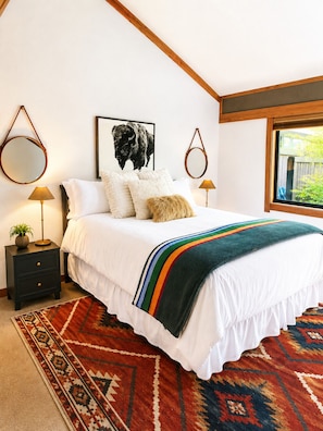 Un dormitorio con una cama, dos mesitas de noche, un espejo, un cuadro enmarcado y una ventana.