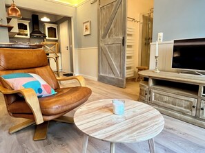 Ein Wohnzimmer mit braunem Ledersessel, einem runden Holztisch, einem Fernseher und einer rustikalen Holzkommode.