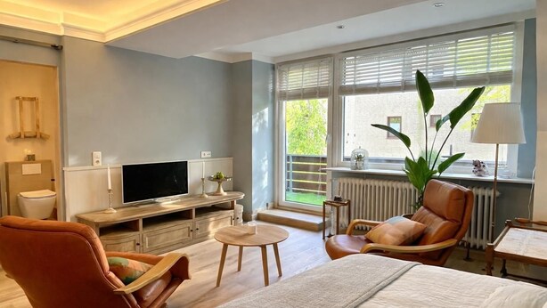 Ein modernes Schlafzimmer mit Bett, Fernseher, Sessel, kleinem Tisch und Fenster mit Jalousien.