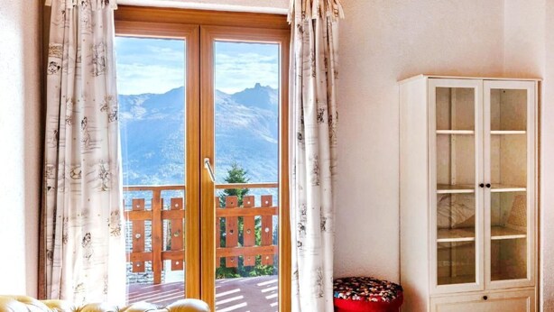 Una habitación con techo de madera, puerta corrediza de vidrio que da a un balcón con vista a la montaña, un reposapiés rojo y un mueble blanco.
