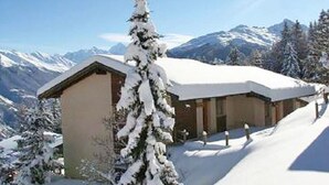 Un albergue cubierto de nieve con montañas al fondo, cielo despejado y un árbol nevado en primer plano.
