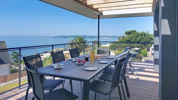 Een overdekt terras met eethoek en uitzicht op zee, een gedekte tafel met borden, glazen en een fles, en stoelen rond de tafel.