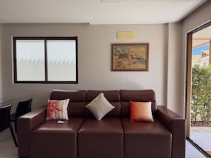 A brown leather sofa with three cushions, a painting of a beach scene, and a window with frosted glass. - Villas de Samoa - Bangalô com 4 quartos (Cabedelo)