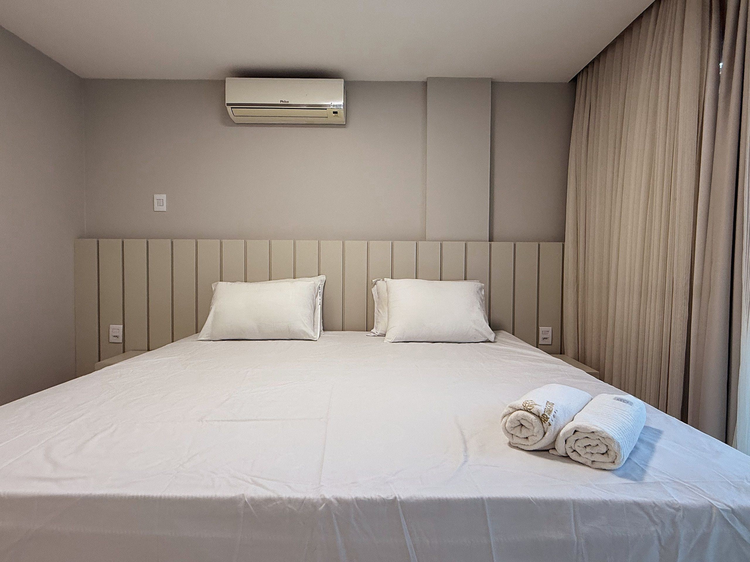 Habitación de hotel con cama, dos almohadas, una toalla enrollada, un equipo de aire acondicionado y una cortina.