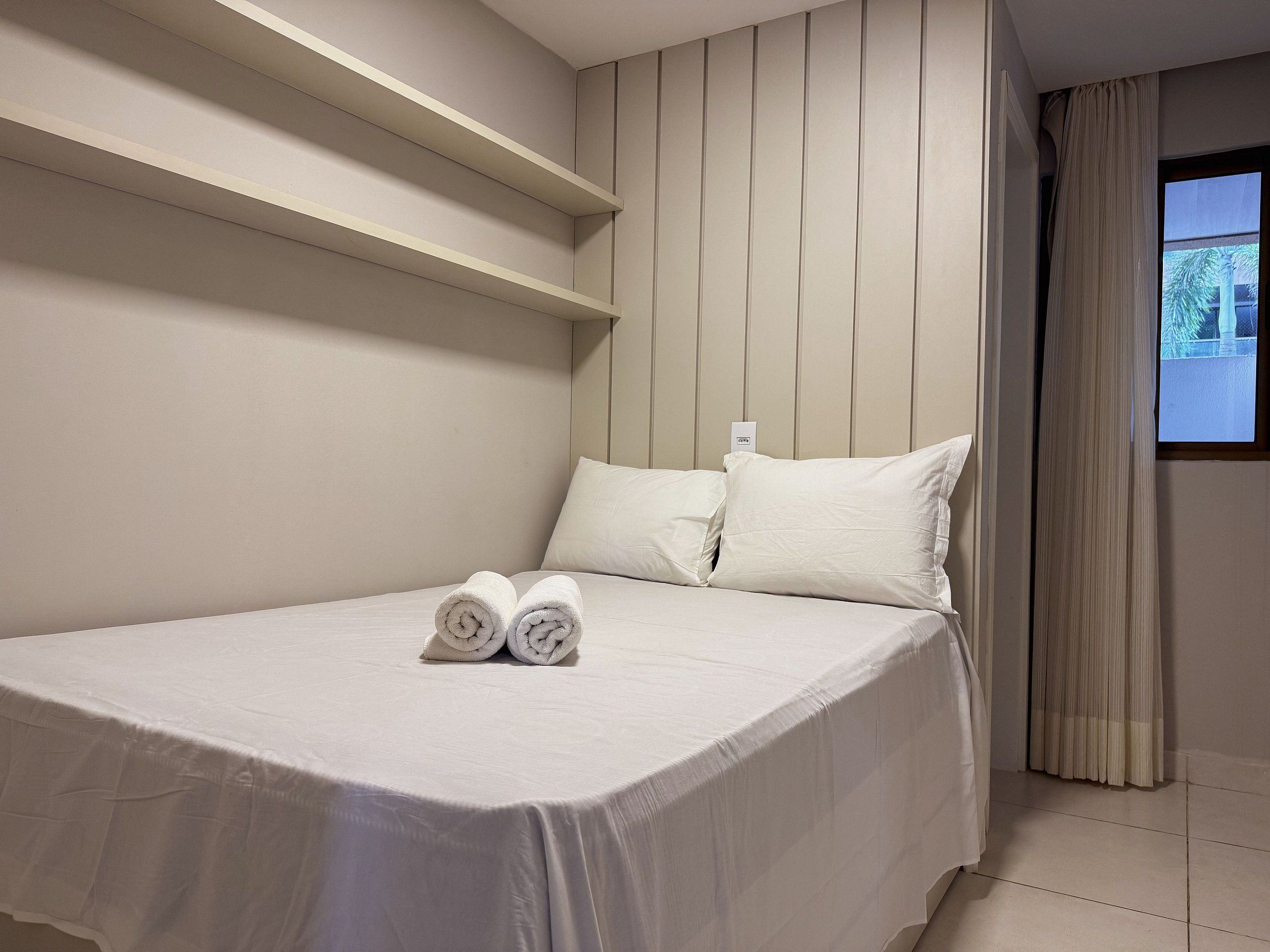 Habitación de hotel con cama, almohadas, una toalla doblada y un ventanal con cortinas.