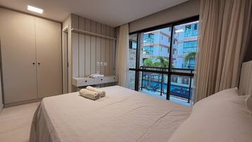 A hotel room with a bed, a wardrobe, a vanity with a mirror, and a large window with a view of a swimming pool and buildings.