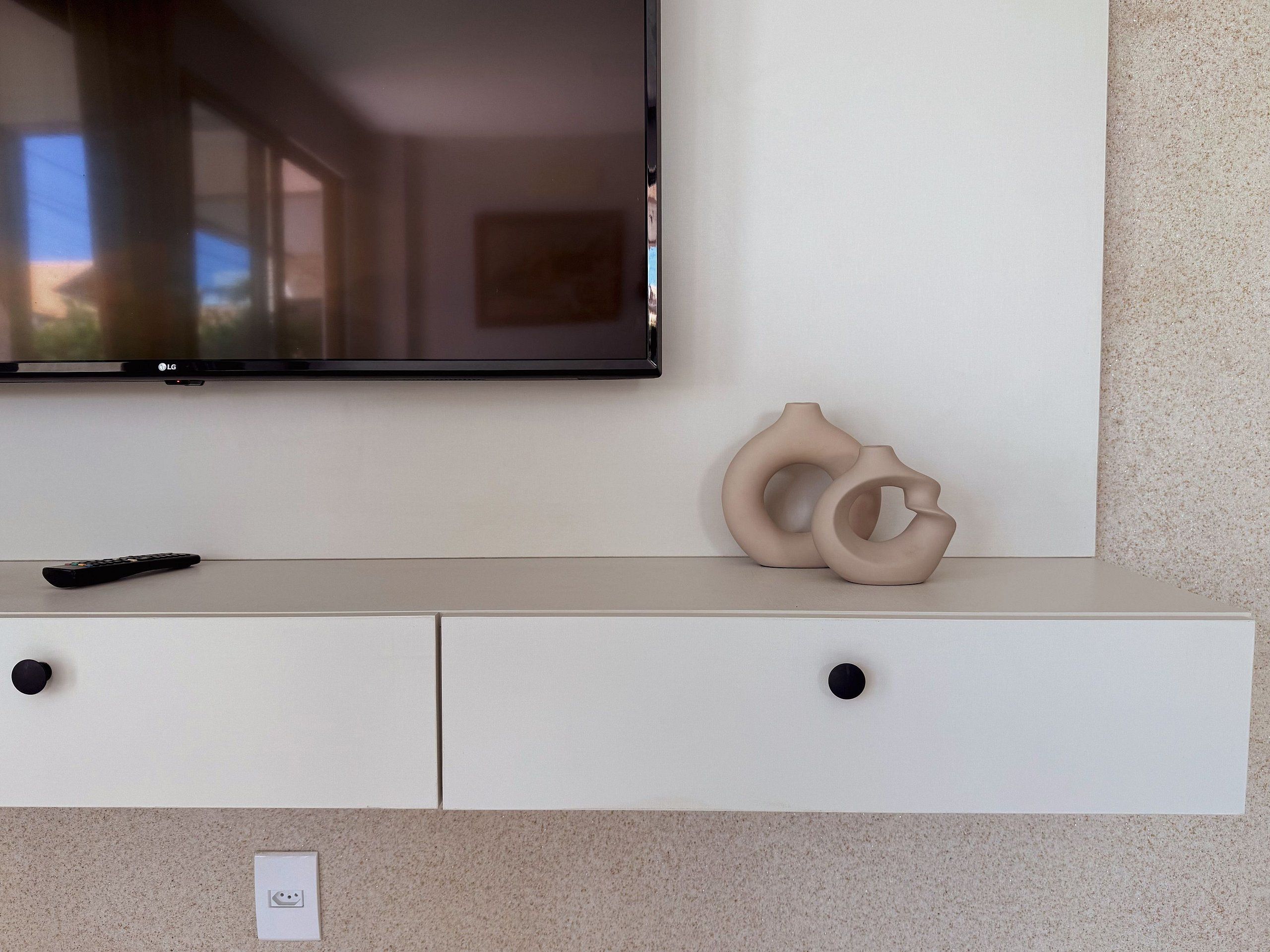 A flat-screen TV mounted on a wall above a white cabinet with two drawers and a remote control on the cabinet's surface.