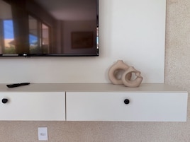 Televisão de tela plana presa na parede acima de um armário branco com duas gavetas e um controle remoto sobre a superfície do armário.