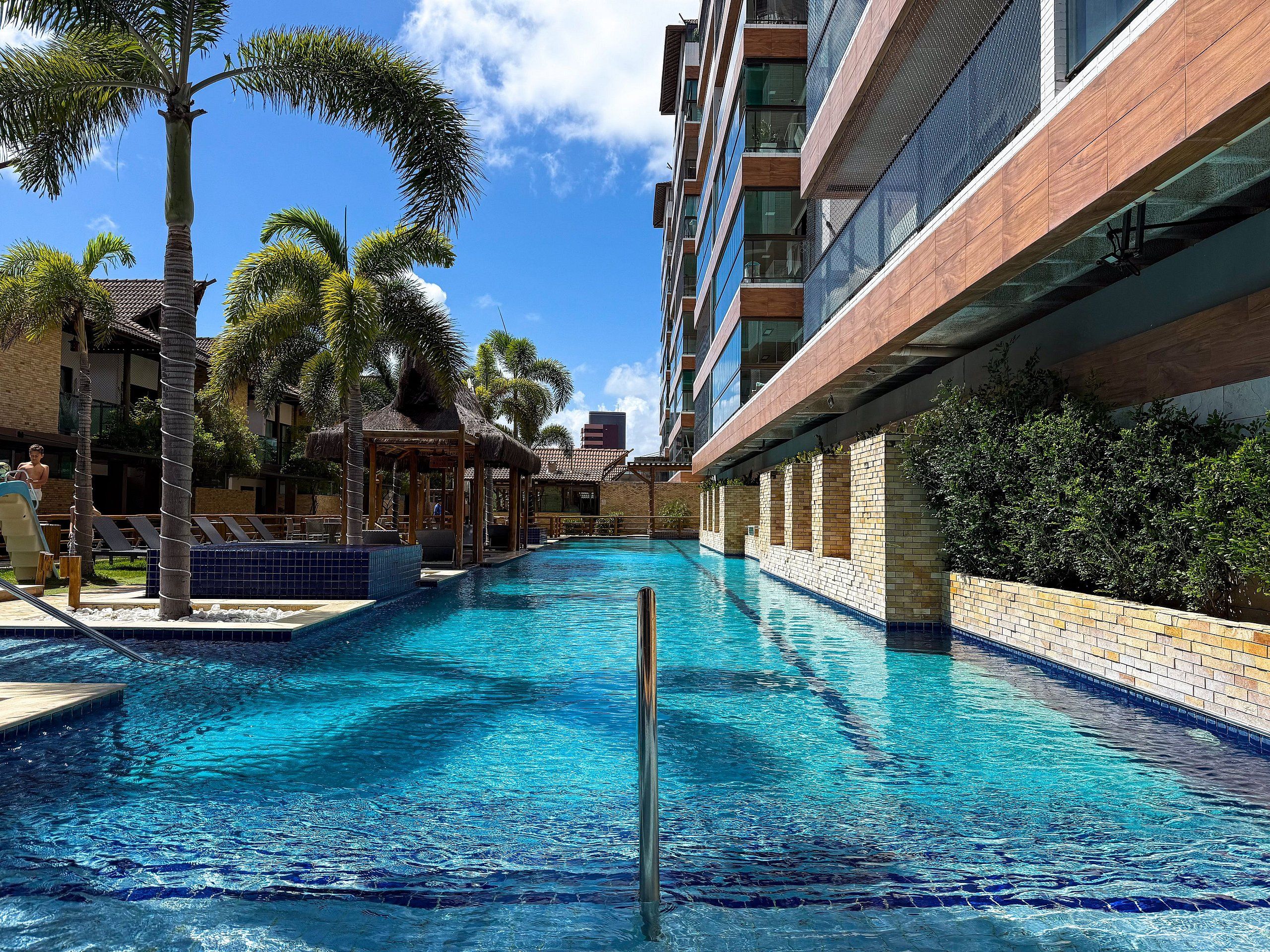 Una piscina con superficie de agua azul cristalina, rodeada de palmeras y un edificio moderno con balcones.
