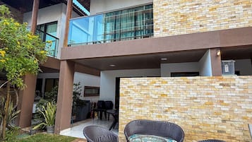 Un edificio de dos pisos con un balcón, zona de estar exterior con sillas de mimbre y una pared de ladrillo.