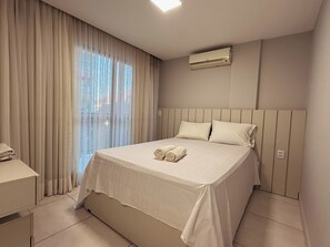 A bedroom with a bed, curtains, an air conditioning unit, and a bedside table.