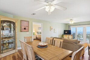 A dining area with a wooden table and chairs, a ceiling fan, and a view of the ocean through large windows. - Amalfi Coast 423c- Ocean Stars & Stripes (Miramar Beach)