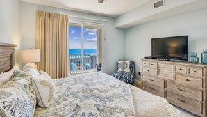 Une chambre avec un lit, un téléviseur posé sur une commode, un ventilateur de plafond et une vue sur l’océan par une porte coulissante.