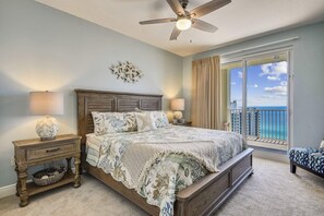 A bedroom with a large bed, a ceiling fan, a nightstand with a lamp, a decorative wall piece, and a view of the ocean through a balcony. - Ariel Dunes I 1909- The Radiant Vista (Miramar Beach)