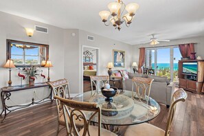 A spacious living room with a glass dining table, comfortable seating, a large window with a view, and a ceiling fan. - Ariel Dunes I 610- Emerald Eden (Miramar Beach)