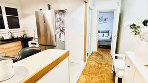 Een smalle gang met aan één kant een keuken, een eethoek met tafel en stoelen, en een slaapkamer zichtbaar door een geopende deur.