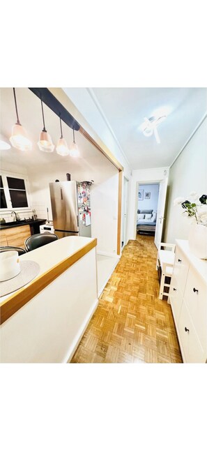 A narrow hallway with a kitchen on one side, a dining area with a table and chairs, and a bedroom visible through an open door. - Elegant 2BR Apartment with Balcony & Free Parking (Ivry-sur-Seine)