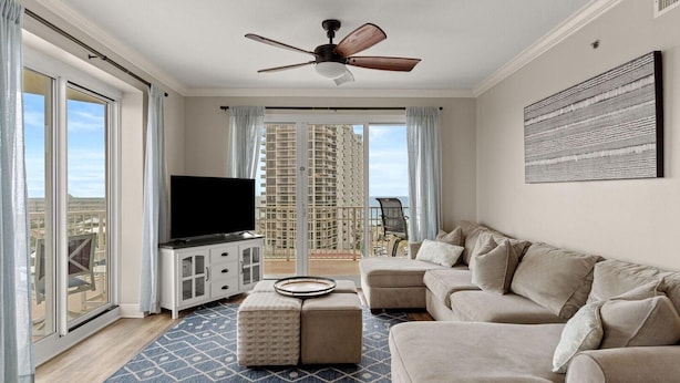 A modern living room with a large flat-screen TV, a beige sectional sofa, a ceiling fan, and a view of high-rise buildings through large windows. - Ariel Dunes I 801- Morning Raes (Miramar Beach)