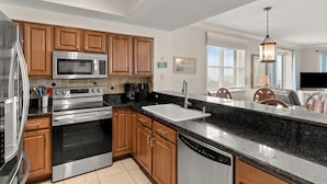 A modern kitchen with stainless steel appliances, wooden cabinets, a black countertop, and a view of the living area. - Ariel Dunes I 801- Morning Raes (Miramar Beach)