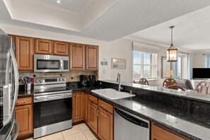 A modern kitchen with stainless steel appliances, wooden cabinets, a black countertop, and a view of the living area. - Ariel Dunes I 801- Morning Raes (Miramar Beach)