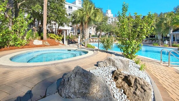 A swimming pool with a wooden deck, palm trees, and a building in the background. - Bahia 4423- Changes in Latitudes (Miramar Beach)