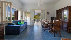 Zona d'estar àmplia amb un sofà blau, taula de dinar i cadires, cuina amb armaris i una vista a un pati exterior a través d'una porta oberta.
