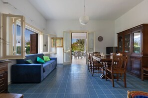 Ein geräumiger Wohnbereich mit blauem Sofa, Esstisch und Stühlen, einer Küchenzeile mit Schränken und Blick durch eine offene Tür auf eine Terrasse.