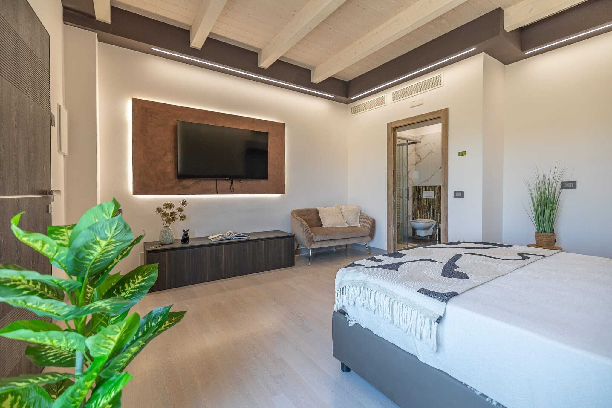 A modern bedroom with a large bed, a flat-screen TV mounted on the wall, a small seating area, and a potted plant.