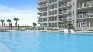 Una piscina con lettini e ombrelloni, un edificio a più piani con balconi e palme.