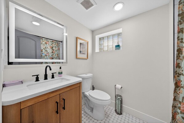 A bathroom with a white sink, wooden cabinets, a toilet, and a small window with a decorative screen. - The Stadium by Mercer Stays | Private 2.2Acre Sports Estate for Teams & Families (Saint Charles)