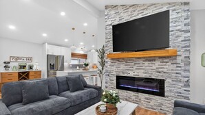 A modern living room with a grey sofa, a marble coffee table, a flat-screen TV mounted on a stone wall, and a built-in fireplace. - The Stadium by Mercer Stays | Private 2.2Acre Sports Estate for Teams & Families (Saint Charles)