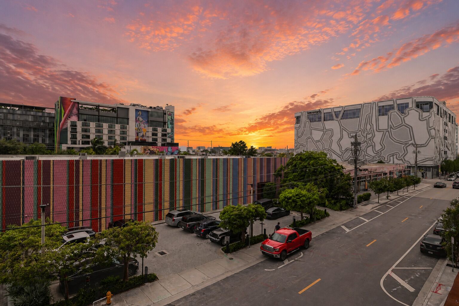 Un edificio moderno con una facciata geometrica particolare, un parcheggio con diverse auto e un cielo al tramonto.